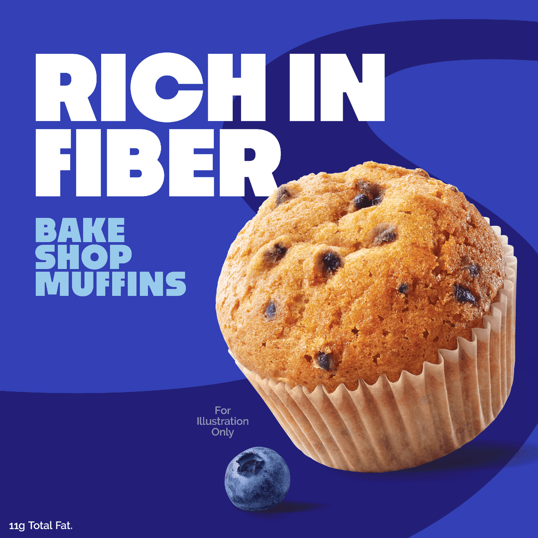 A blueberry muffin in a paper liner cup on a blue graphic background with large white text reading 'RICH IN FIBER' and smaller text 'BAKE SHOP MUFFINS'. A single blueberry is shown near the bottom left, with small text 'For Illustration Only' and '11g Total Fat.