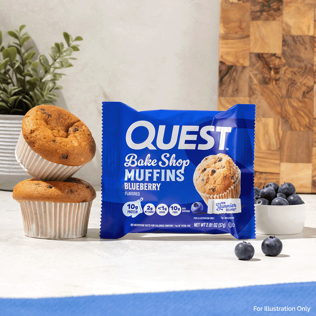 A package of Quest Bake Shop Blueberry Muffins stands on a countertop, flanked by two baked muffins, a small bowl of blueberries, and a wood cutting board in the background.