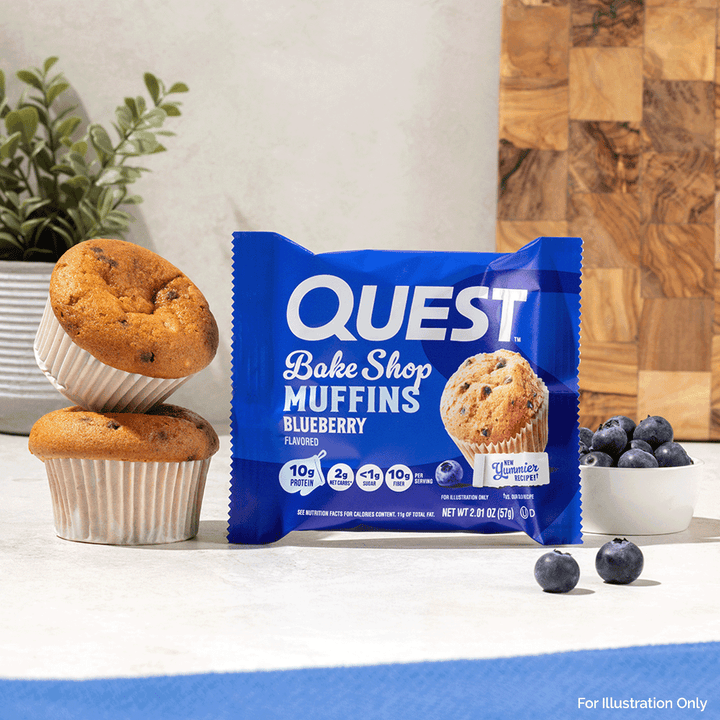 A package of Quest Bake Shop Blueberry Muffins stands on a countertop, flanked by two baked muffins, a small bowl of blueberries, and a wood cutting board in the background.