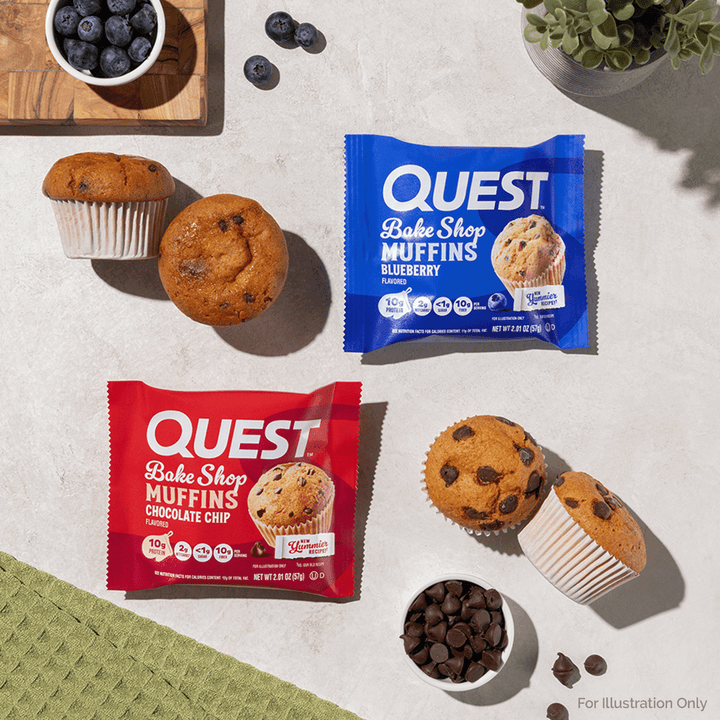 Two Quest Bake Shop Muffins snack bars, one blueberry and one chocolate chip, arranged on a light stone surface with muffins, chocolate chips, blueberries, and a small plant.
