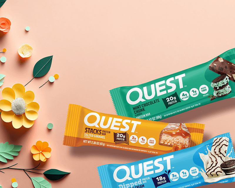Three Quest protein bars in colorful wrappers, mint chocolate, salted caramel, and dipped ice cream, are arranged on a peach background with paper flowers.