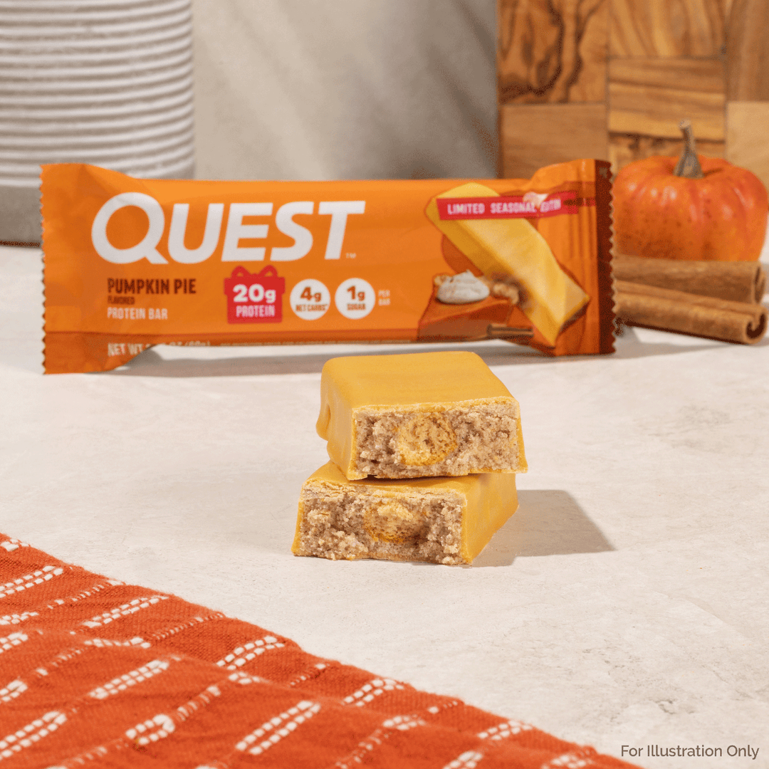 Two Quest Pumpkin Pie protein bars stacked on a light surface, with the orange Quest packaging blurred in the background and a small pumpkin and cinnamon sticks behind it.