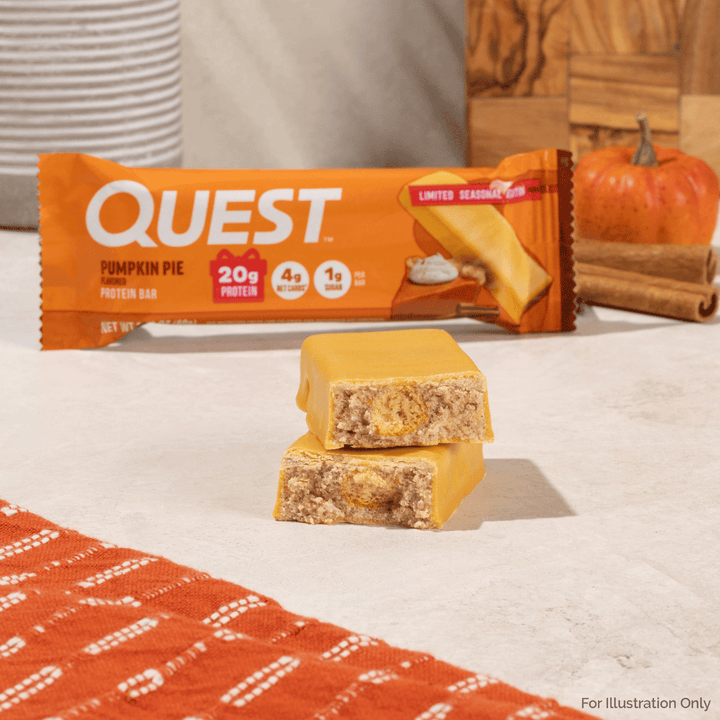 Two Quest Pumpkin Pie protein bars stacked on a light surface, with the orange Quest packaging blurred in the background and a small pumpkin and cinnamon sticks behind it.