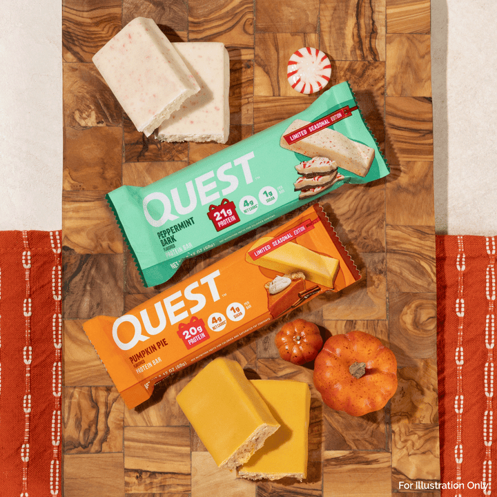 Two Quest snack bars, one Peppermint Milk Chocolate and one Pumpkin Pie Chocolate, arranged on a wooden board with candy canes, small pumpkins, and cheese slices. The Quest packaging is visible, and the scene is styled with autumn-themed props.
