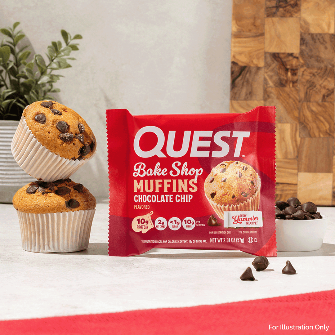 A package of Quest Bake Shop Muffins Chocolate Chip is displayed on a countertop with two chocolate chip muffins, chocolate chips, and a wooden cutting board in the background.