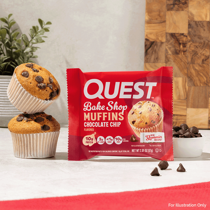 A package of Quest Bake Shop Muffins Chocolate Chip is displayed on a countertop with two chocolate chip muffins, chocolate chips, and a wooden cutting board in the background.