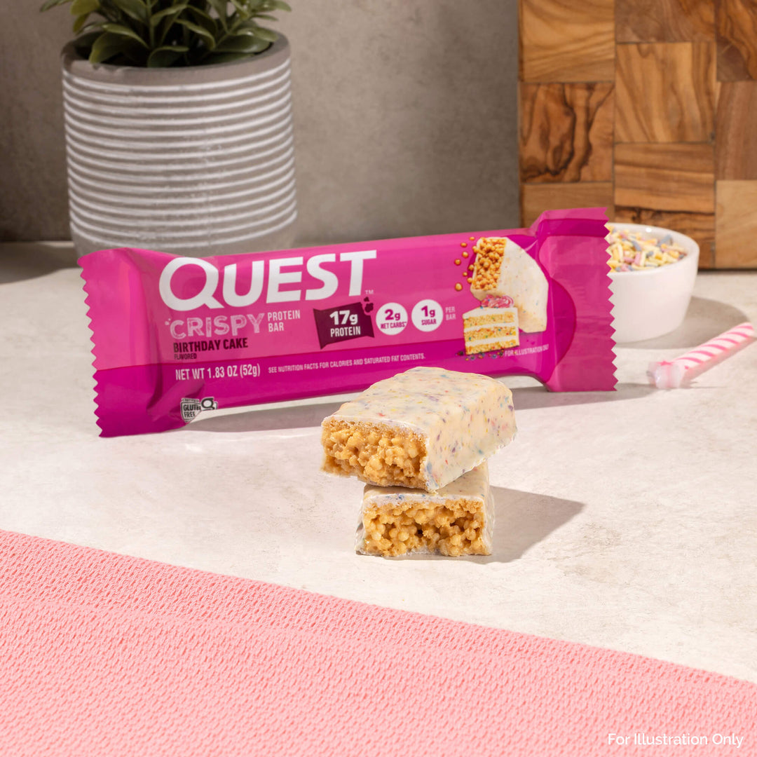 Quest Crispy Protein Bar in Birthday Cake flavor on a pink surface with a plant and wooden cutting board in the background.