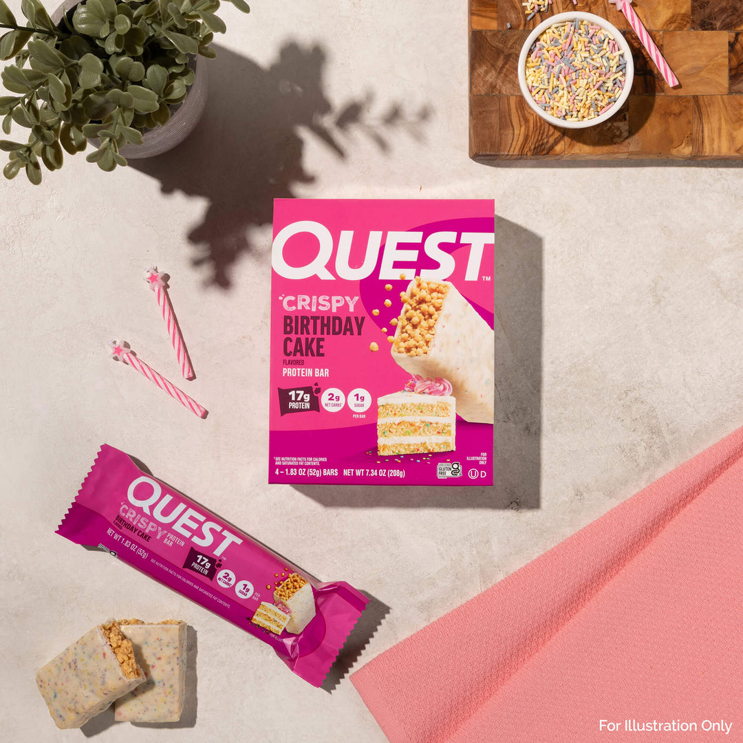 Quest Crispy Birthday Cake protein bar packaging on a light surface with a plant and pink napkin.