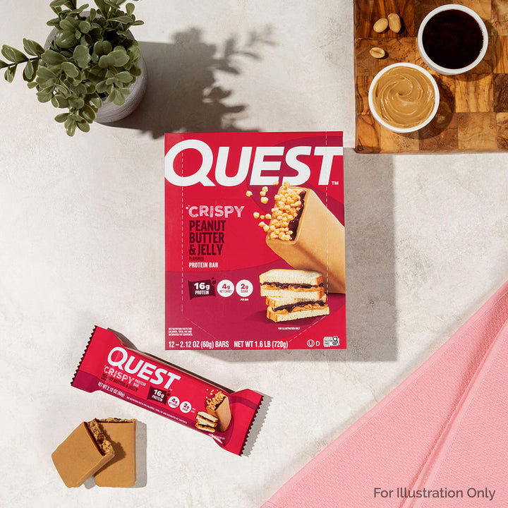 Quest Crispy Peanut Butter & Jelly protein bar packaging on a light surface with a plant and coffee cup in the background.