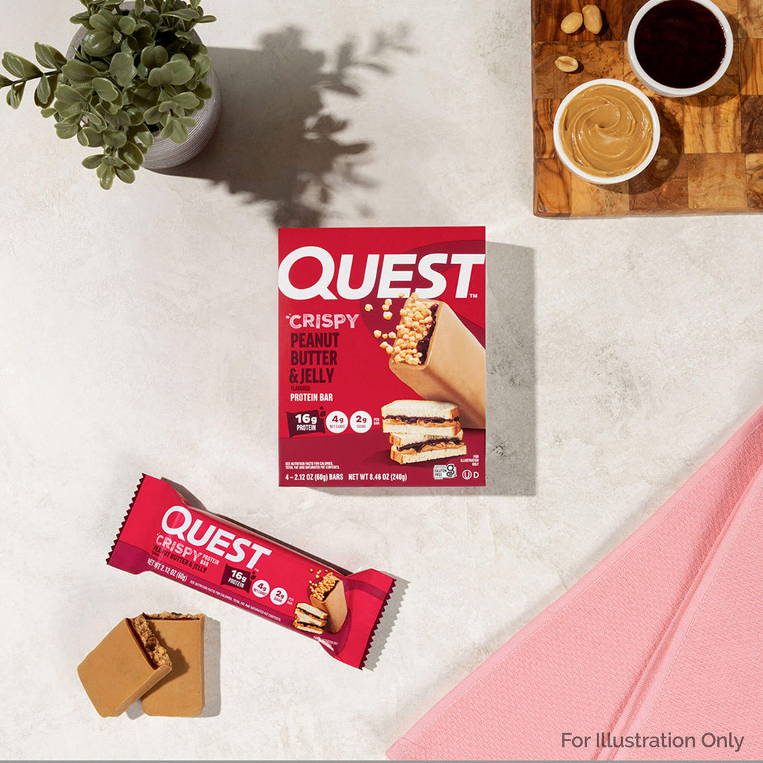 Quest Crispy Peanut Butter & Jelly protein bar packaging on a light surface with a plant and coffee cup in the background.