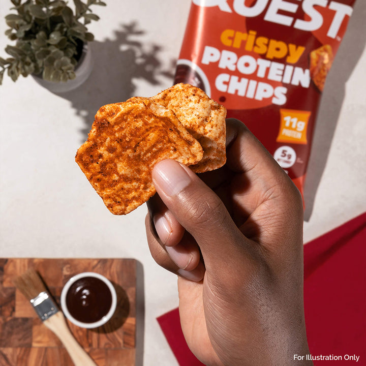 Hand holding crispy protein chips near a BBQ sauce cup and brush on a wooden board. A red chip bag and potted plant in the background.