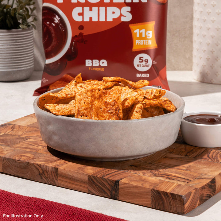 A bowl of BBQ-flavored protein chips is on a wooden board. Behind it, a bag of chips displays "11g Protein." A cup of dip sits beside the bowl.