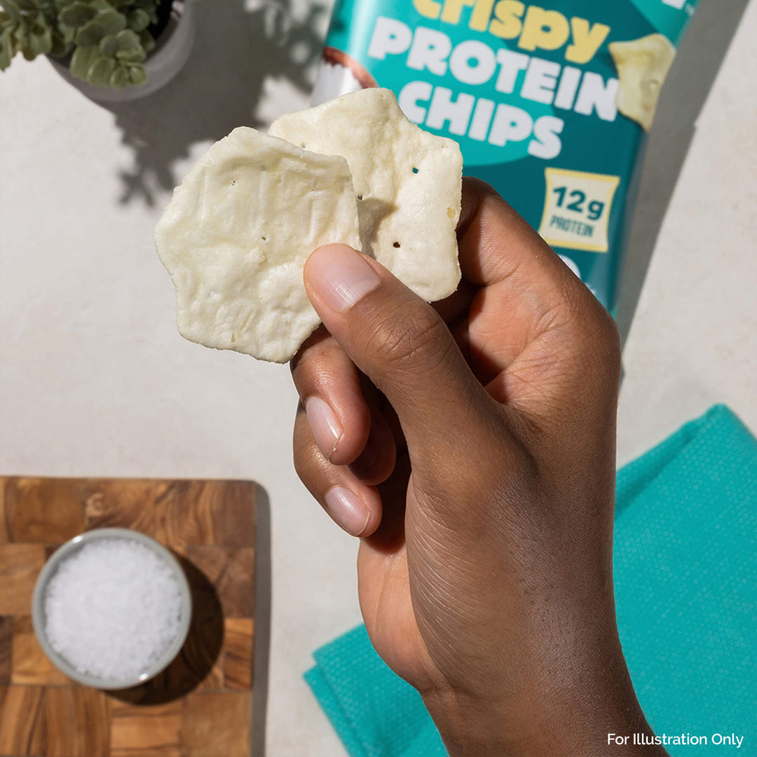 Hand holding two white, hexagonal protein chips above a wooden board with a bowl of salt. A teal bag labeled "Crispy Protein Chips" is partially visible.