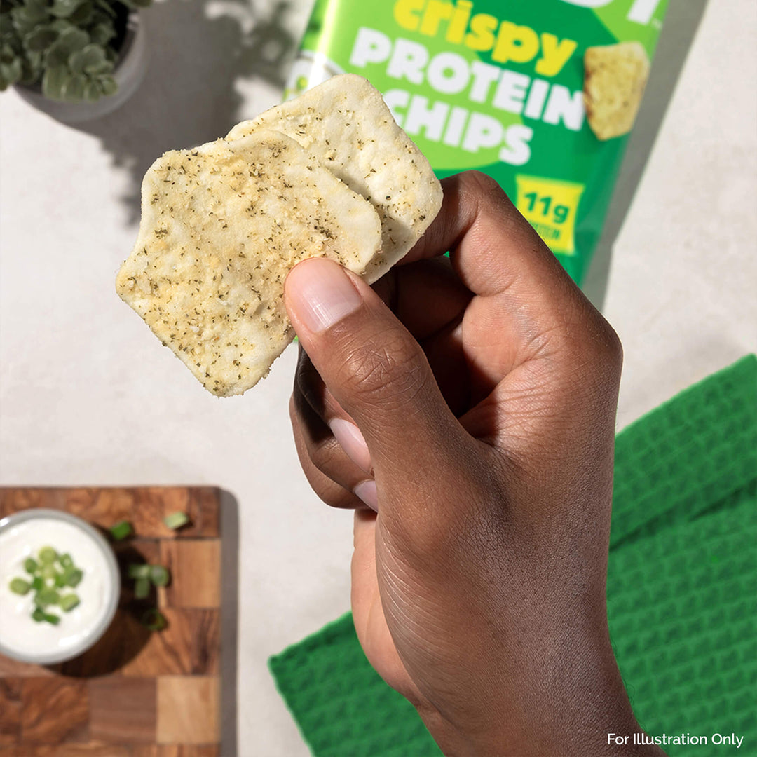 A hand holds two seasoned, square protein chips. In the background, a green chip bag reads "crispy protein chips." A cutting board with a dip bowl and diced scallions is nearby.