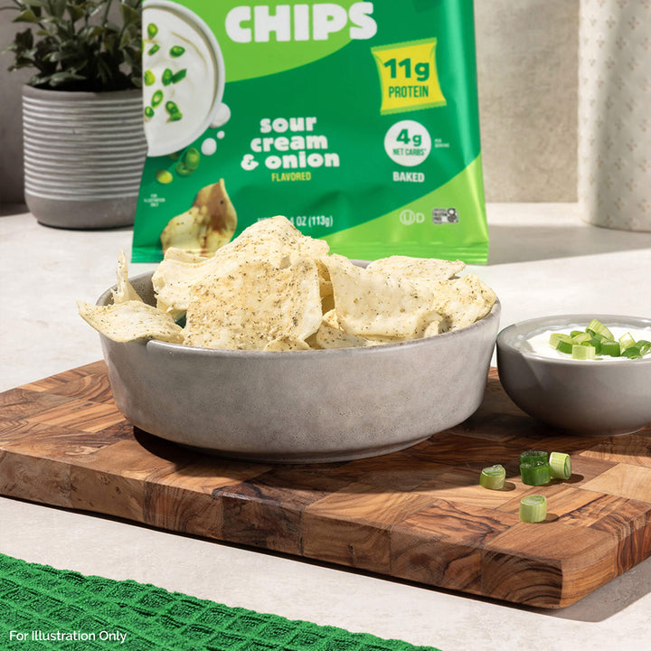 A bowl of sour cream and onion chips sits on a wooden board beside a small dish of dip topped with chopped green onions. A green chip bag is in the background.