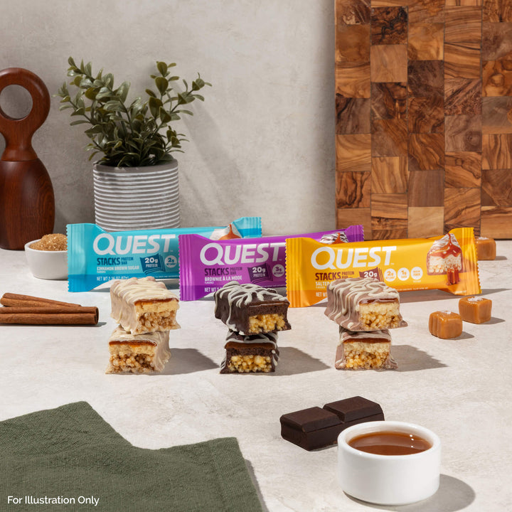 Assorted Quest protein bars are stacked near ingredients like caramel, cinnamon, and chocolate on a light kitchen countertop, conveying a tasty, inviting scene.