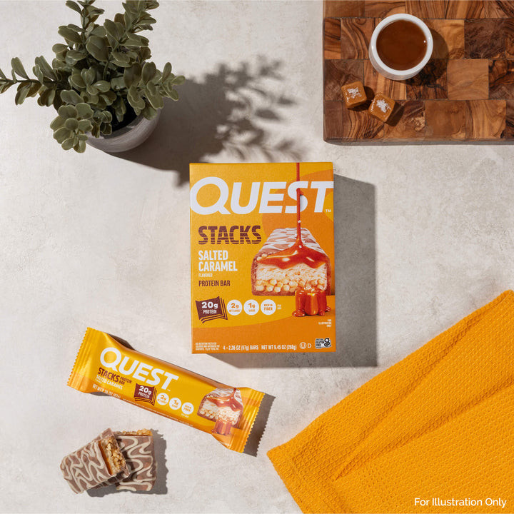 Box of Quest Stacks Salted Caramel protein bars next to an unwrapped bar, caramel bowl, and yellow towel on a light stone surface.
