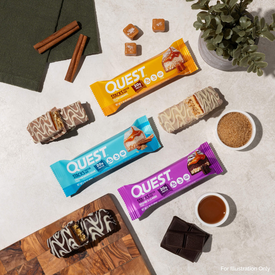 Three Quest protein bars with caramel, s’mores, and chocolate themes are on a table with cinnamon sticks, sugar, chocolate, caramel pieces, and a plant.