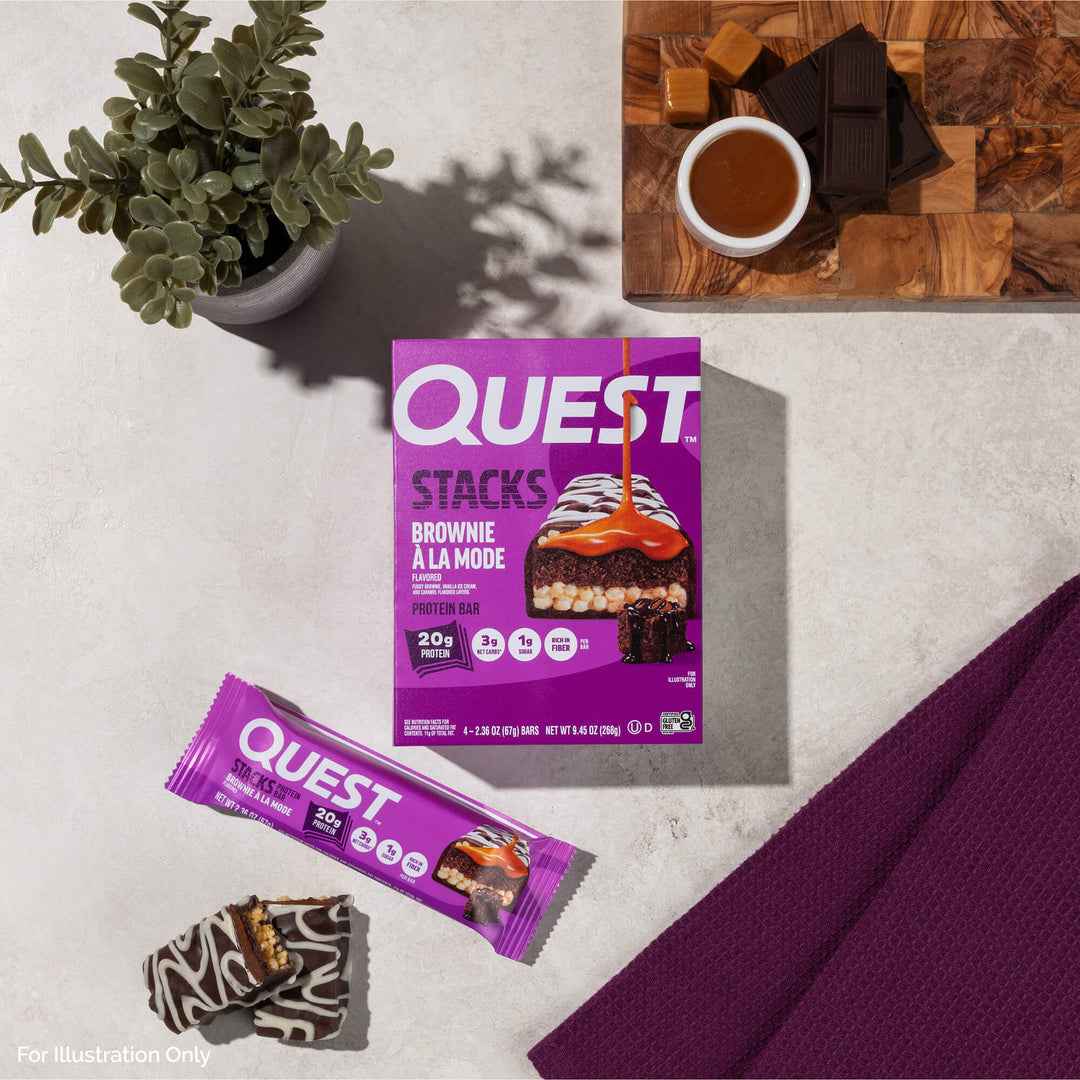 Box of Quest Stacks Brownie À La Mode protein bars on a light surface. Nearby, a tray with chocolate, caramel, a purple bar wrapper, and a plant.