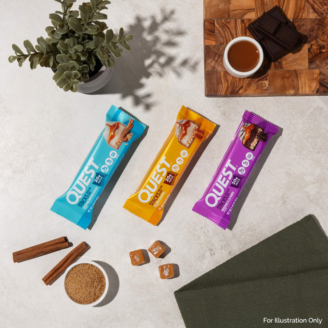 Three Quest protein bars are arranged on a light surface with a plant, cinnamon sticks, sugar, caramel pieces, and chocolate on a wooden board.