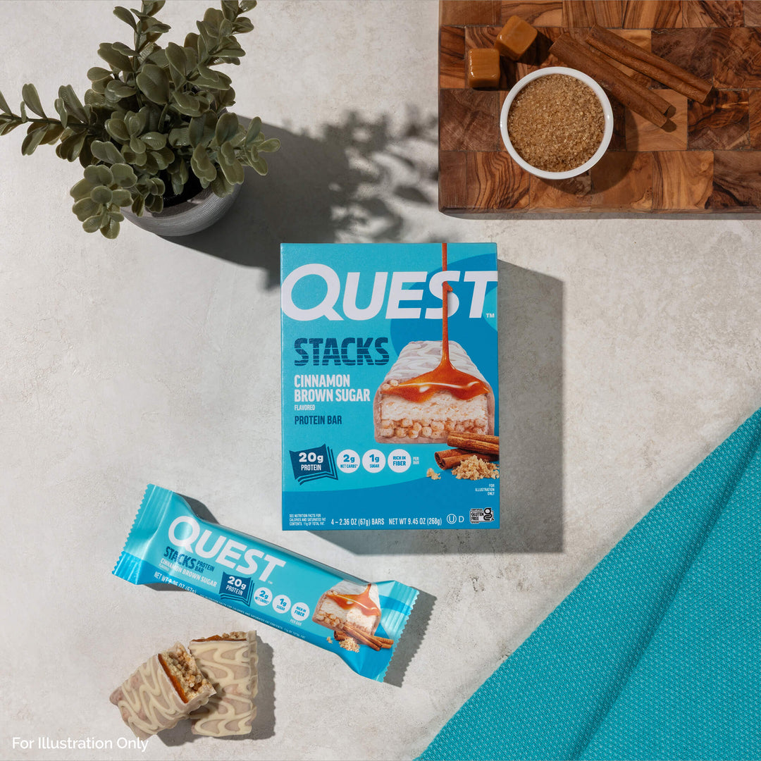 Box and wrapper of Quest Cinnamon Brown Sugar protein bars on a light surface. Nearby are caramel cubes, brown sugar, cinnamon sticks, and a plant.