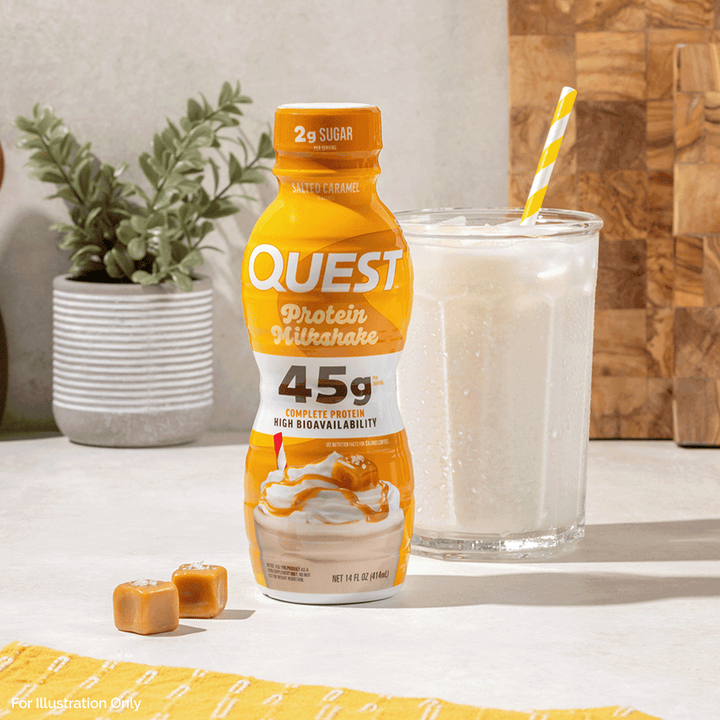 A bottle of Quest Protein Ultra Shake is shown on a countertop beside a glass of creamy beverage with a yellow-and-white striped straw, with two small caramel pieces in front and a potted plant and wood paneling in the background.